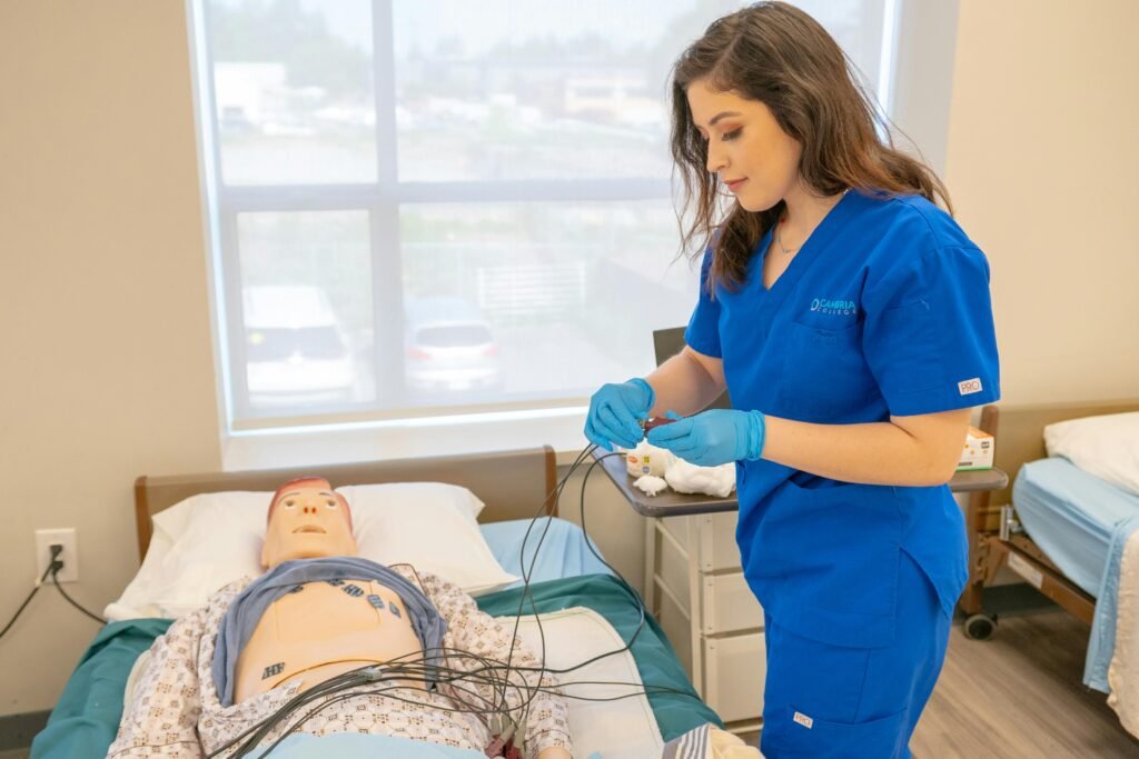 Medical student in blue scrubs practicing procedures on a patient training manikin in a clinical setting.