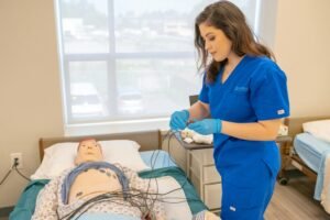 Medical student in blue scrubs practicing procedures on a patient training manikin in a clinical setting.